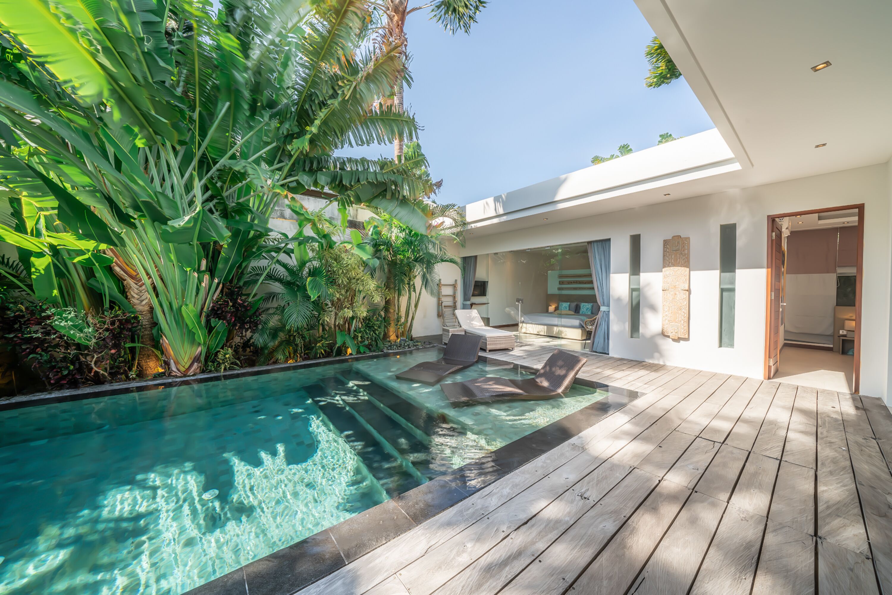 Un área de piscina moderna al aire libre con cubierta de madera, sillones de descanso y exuberante vegetación tropical.
