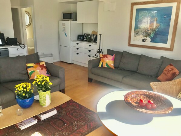 Un salon avec deux canapés gris, une table basse et une table à manger agrémentée d’un vase décoratif. On aperçoit également un coin cuisine avec un four à micro-ondes et un réfrigérateur au fond.