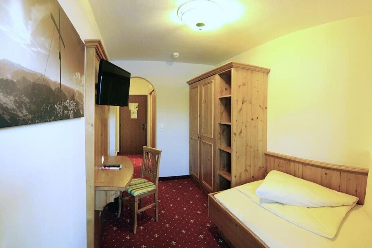 Ein kleiner, gemütlicher Raum mit einem Bett, einem Schreibtisch und einem Stuhl. Es gibt einen Holzschrank und ein Bild an der Wand.