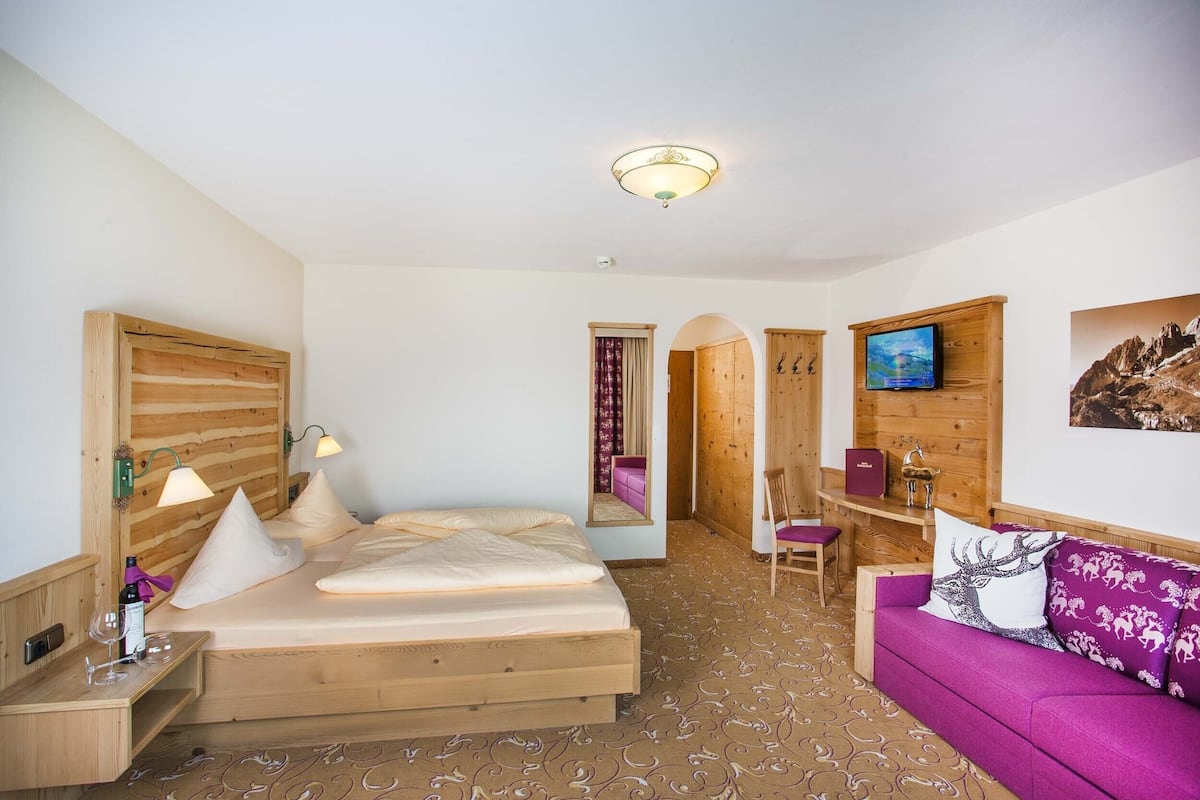 Ein Bett mit Holzkopfteil, ein Schreibtisch mit Stuhl, ein violettes Sofa und ein Fernseher an der Wand.