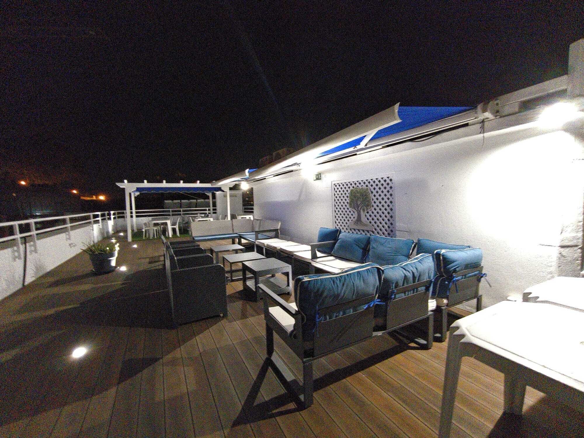 Eine Terrasse im Freien mit Sitzgelegenheiten und Tischen, eine weiße Wand mit einer Baummalerei und einem Geländer.