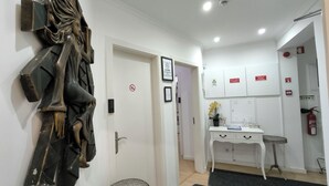 Un pasillo con una escultura, una puerta con un cartel de no fumar, una pequeña mesa redonda con una planta en maceta y un armario blanco con etiquetas rojas.