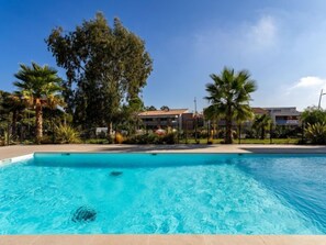 Uma piscina com água azul cristalina, rodeada por palmeiras e um edifício com telhado de azulejos ao fundo.
