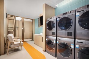 A laundry room with stacked front-loading washers and dryers, a seating area with chairs, and a visible exit door. - Home2 Suites by Hilton Shenyang Yuhong (Shenyang)