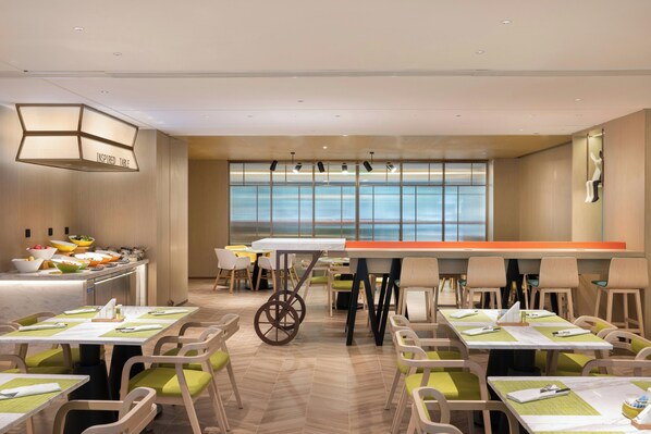 Un comedor moderno con varias mesas y sillas, un mostrador tipo bufé y amplias ventanas que ofrecen luz natural.