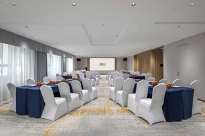 A conference room with rows of tables and chairs, a projector screen, and a large window with curtains. - Home2 Suites by Hilton Shenyang Yuhong (Shenyang)