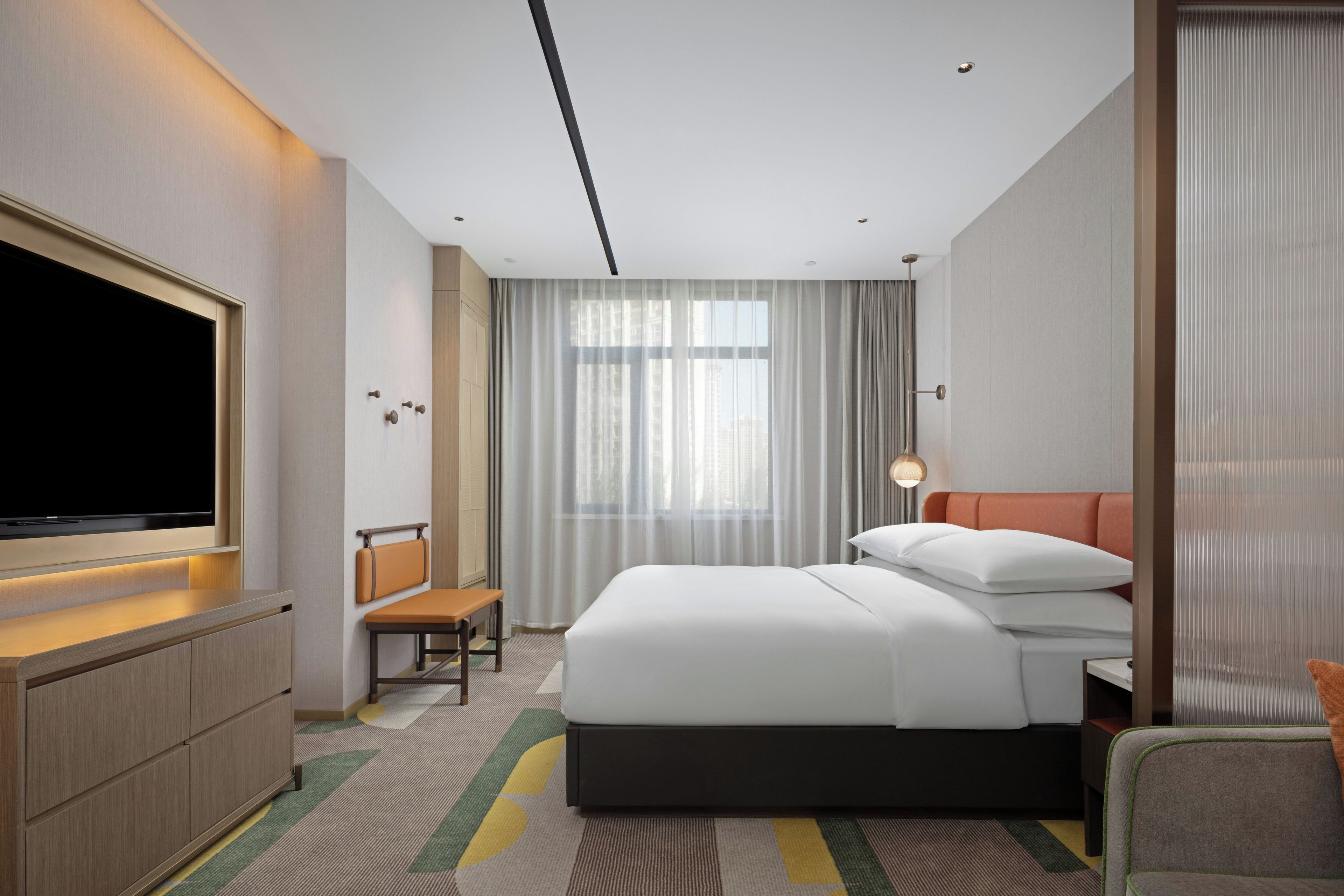 Habitación de hotel con televisor de pantalla plana grande, cama con ropa blanca, escritorio con silla y ventana con cortinas traslúcidas.