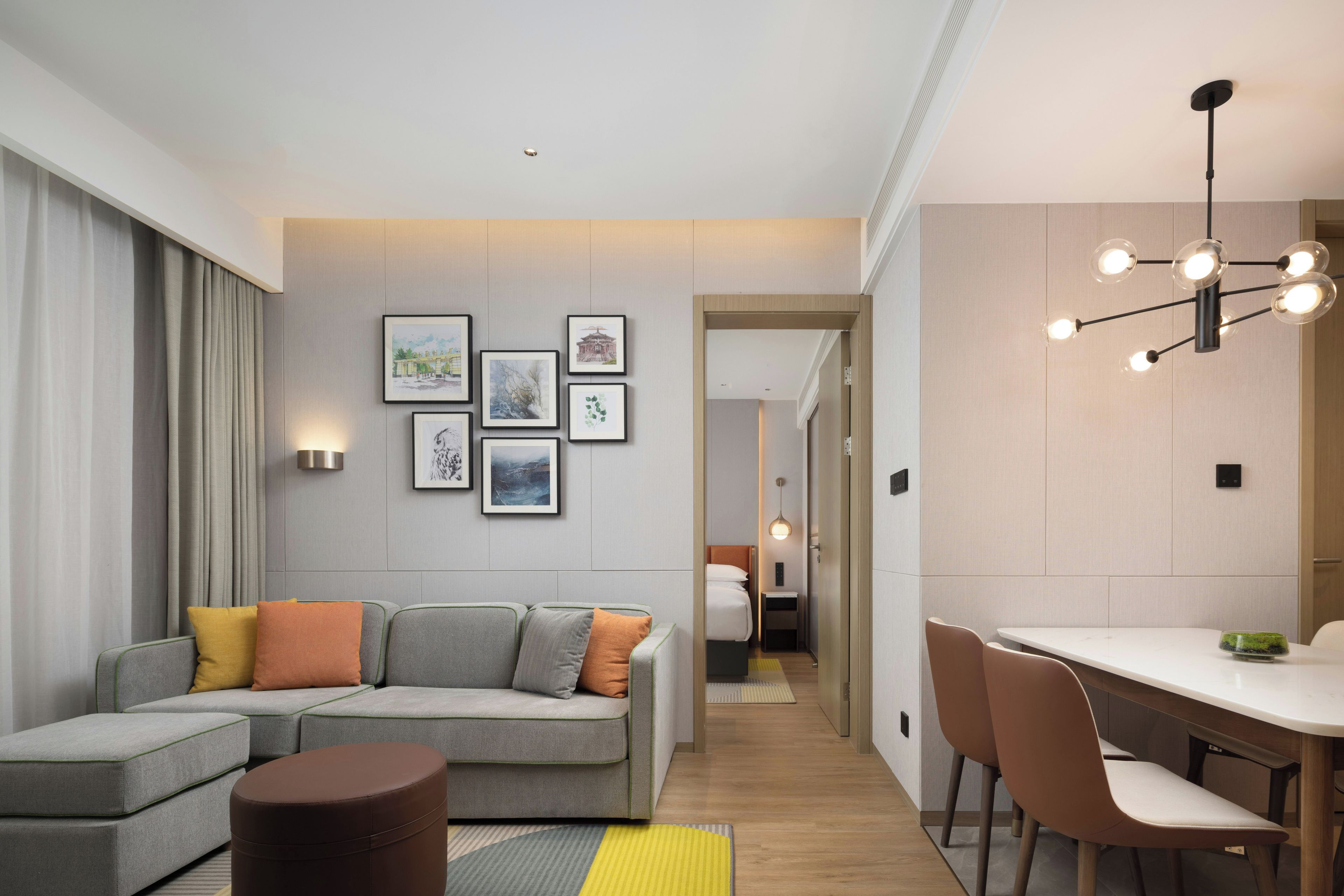 A modern hotel room with a sofa, ottoman, dining table, chairs, and a hallway leading to a bedroom.