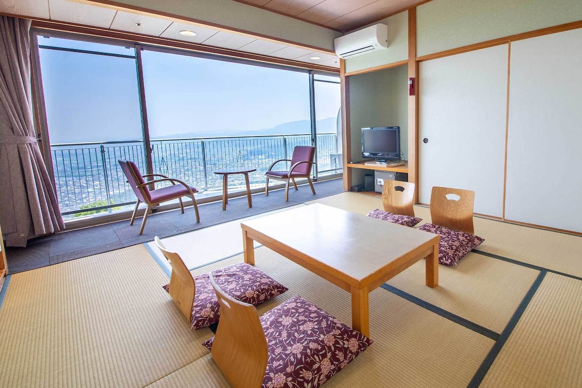 Een traditionele Japanse kamer met tatami vloerbedekking, een lage tafel en zitplaatsen. Een televisie staat op een plank, en er is een groot raam met uitzicht op de stad.
