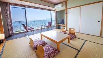 A traditional Japanese room with tatami flooring, a low table, and seating. A television is on a shelf, and there's a large window with a view of the city.