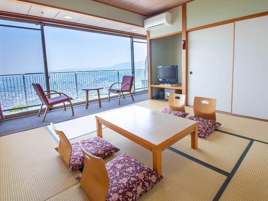 Il s’agit d’une pièce de style japonais traditionnel, avec un sol recouvert de tatamis, une table basse et des sièges. Un téléviseur est posé sur une étagère, et une grande fenêtre offre une vue sur la ville.