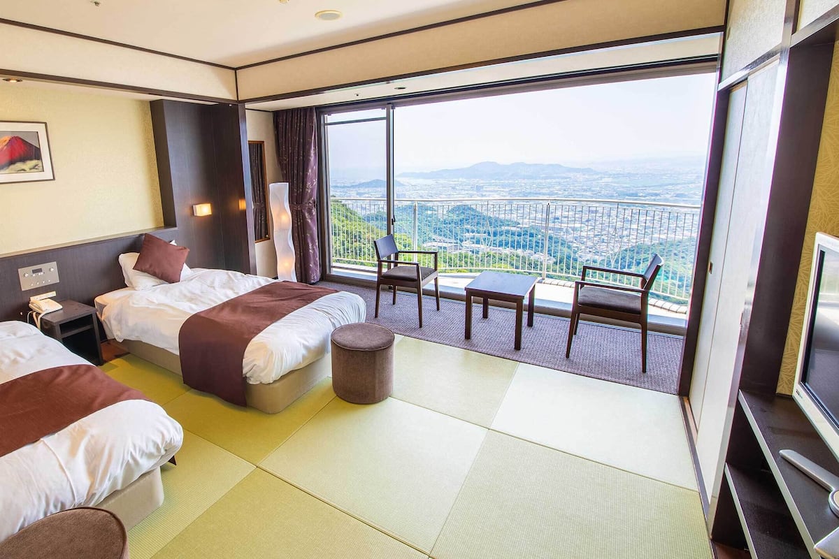 Hotelkamer met twee bedden, een tafeltje, een stoel, een televisie en een balkon met uitzicht over de stad.