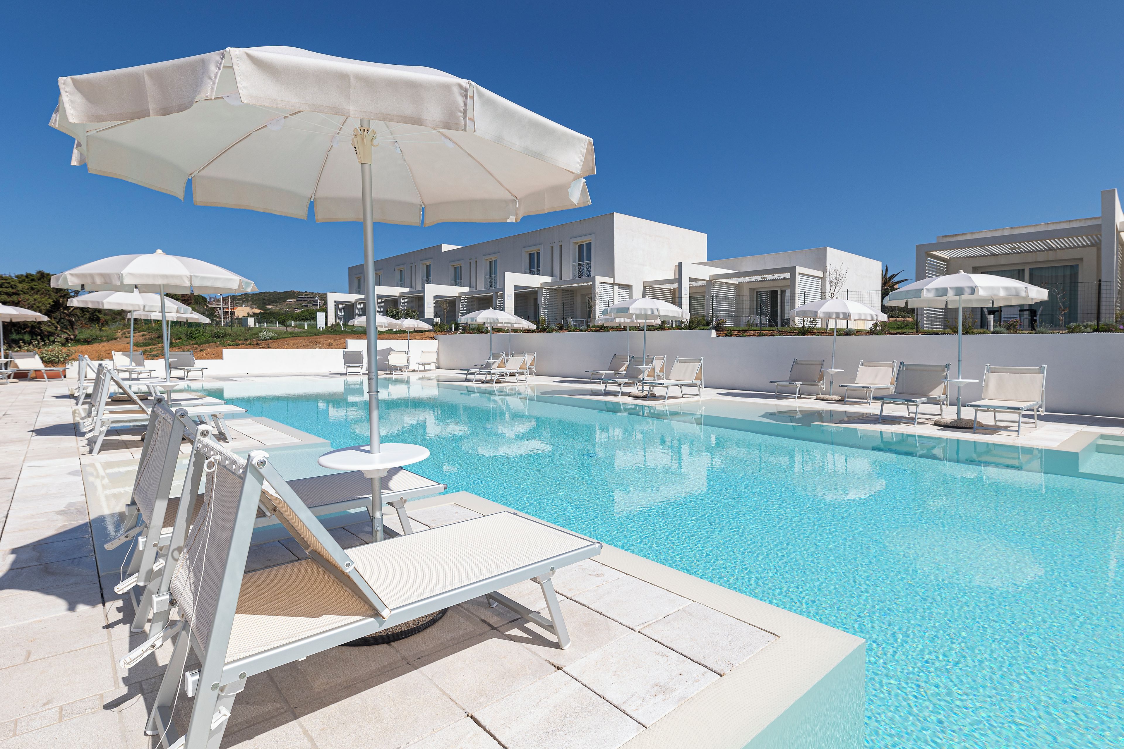 Un área junto a la piscina con sillas reclinables blancas y sombrillas, una piscina azul cristalina y edificios modernos al fondo.
