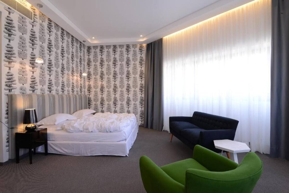 Habitació d'hotel amb un llit, un otomà verd, un sofà negre, una taula petita blanca i un paper pintat amb motius.