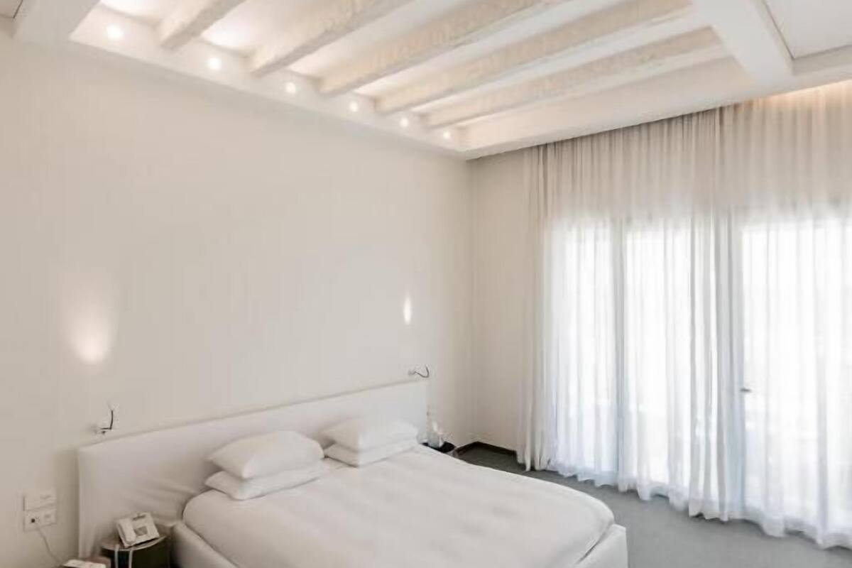 Un dormitori modern amb un llit gran, una taula de nit amb un telèfon i una finestra amb cortines transparents.