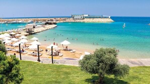 A beach with clear turquoise water, sun loungers, umbrellas, and a modern building in the background. - Miramar Hotel Resort & Spa (Qalamoun)