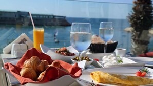 A table with croissants, a glass of water, a glass of white wine, a glass of orange juice, and a plate with a fish dish, overlooking a body of water. - Miramar Hotel Resort & Spa (Qalamoun)