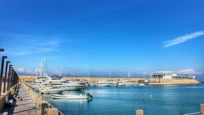A marina with boats docked along a concrete quay, a sandy beach in the background, and a modern building on an island. - Miramar Hotel Resort & Spa (Qalamoun)