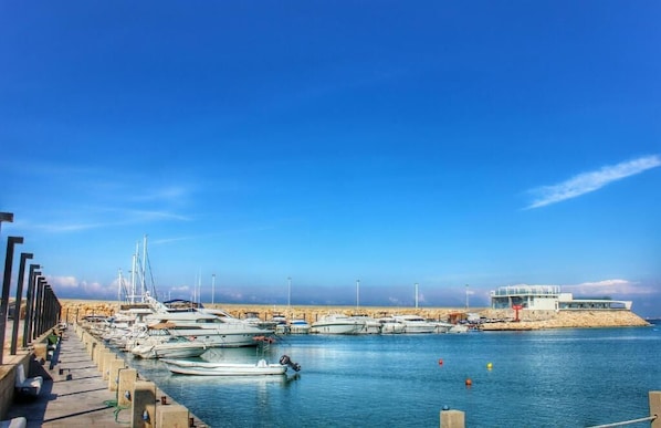 Una marina con barcos amarrados a un muelle de hormigón, una playa de arena al fondo y un edificio moderno en una isla.
