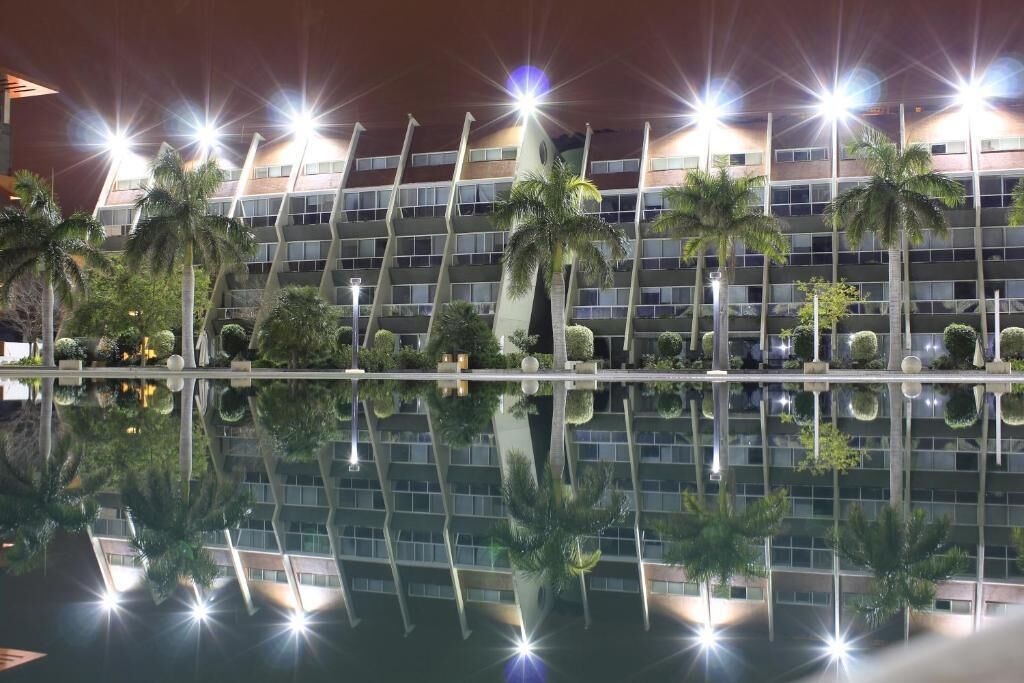 Um hotel com vários andares e design moderno, com uma piscina espelhada em frente e palmeiras ao redor do edifício.
