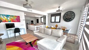 Una sala de estar moderna con un sofá blanco, un tapete colorido, una mesa de centro de madera y un comedor con un reloj en la pared.