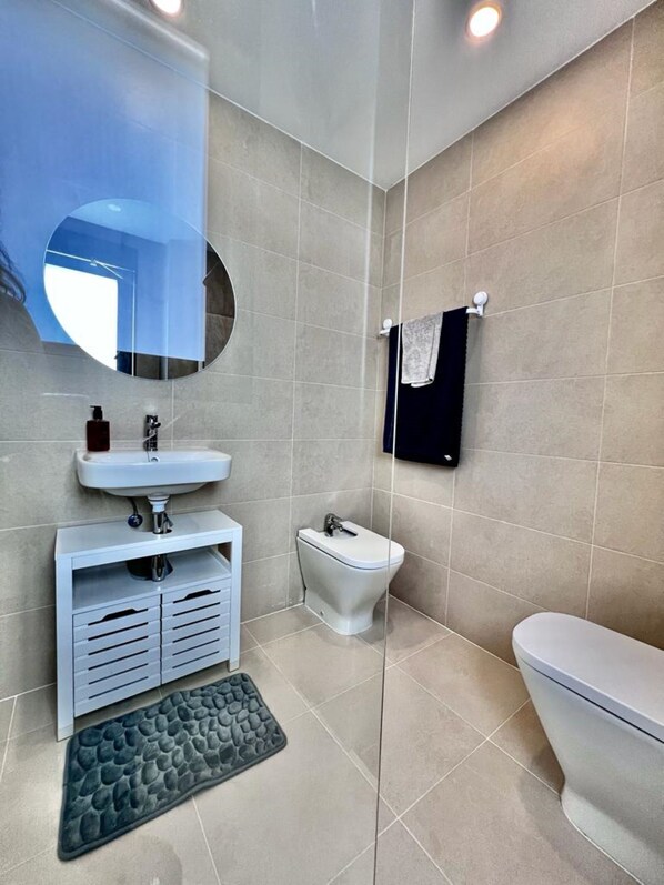 Un baño moderno con un lavamanos blanco, un inodoro y un bidé. Hay un espejo sobre el lavamanos y un perchero en la pared.