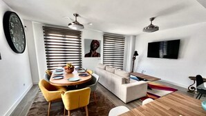 Un salón moderno con una mesa redonda de comedor, sillas amarillas, un sofá color beige y un televisor de pantalla plana montado en la pared.