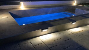 Una piscina exterior moderna con fuente de agua azul, rodeada de baldosas de piedra e iluminada por luces empotradas.