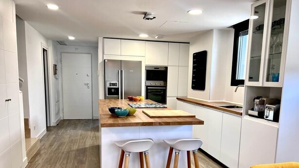 Una cocina moderna con armarios blancos, una isla de madera con taburetes y una silla amarilla.