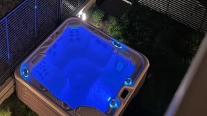 Un jacuzzi iluminado con luz azul, rodeado por una terraza de madera y una valla metálica.