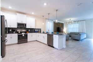 A modern kitchen with white cabinets, a microwave, an oven, and a dishwasher. There is a dining area with a grey sofa and a flat-screen TV. - South Facing Pool! Private Game Room+Near Disney (Kissimmee)