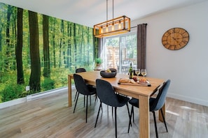 A dining area with a wooden table, black chairs, a large wall mural of a forest, a hanging light fixture, and a wooden clock on the wall.
