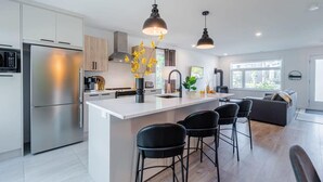 A modern kitchen with a central island, stainless steel appliances, pendant lighting, and a seating area with black chairs. - Le Taiga - Pool Table, Nature, Pet Friendly (Pont-Rouge)