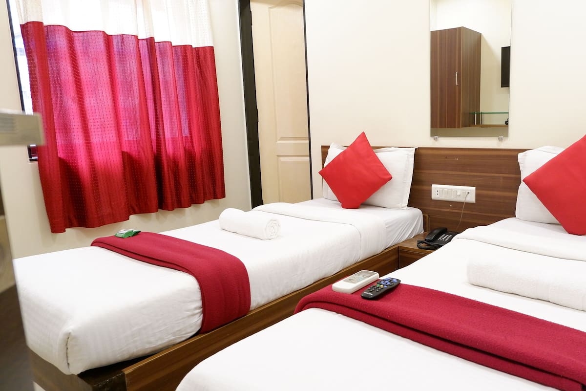 Habitación de hotel con dos camas individuales, cada una con una almohada roja, ropa de cama blanca y un plaid rojo. Tiene un cabecero de madera, una mesita de noche con un teléfono y un espejo en la pared.