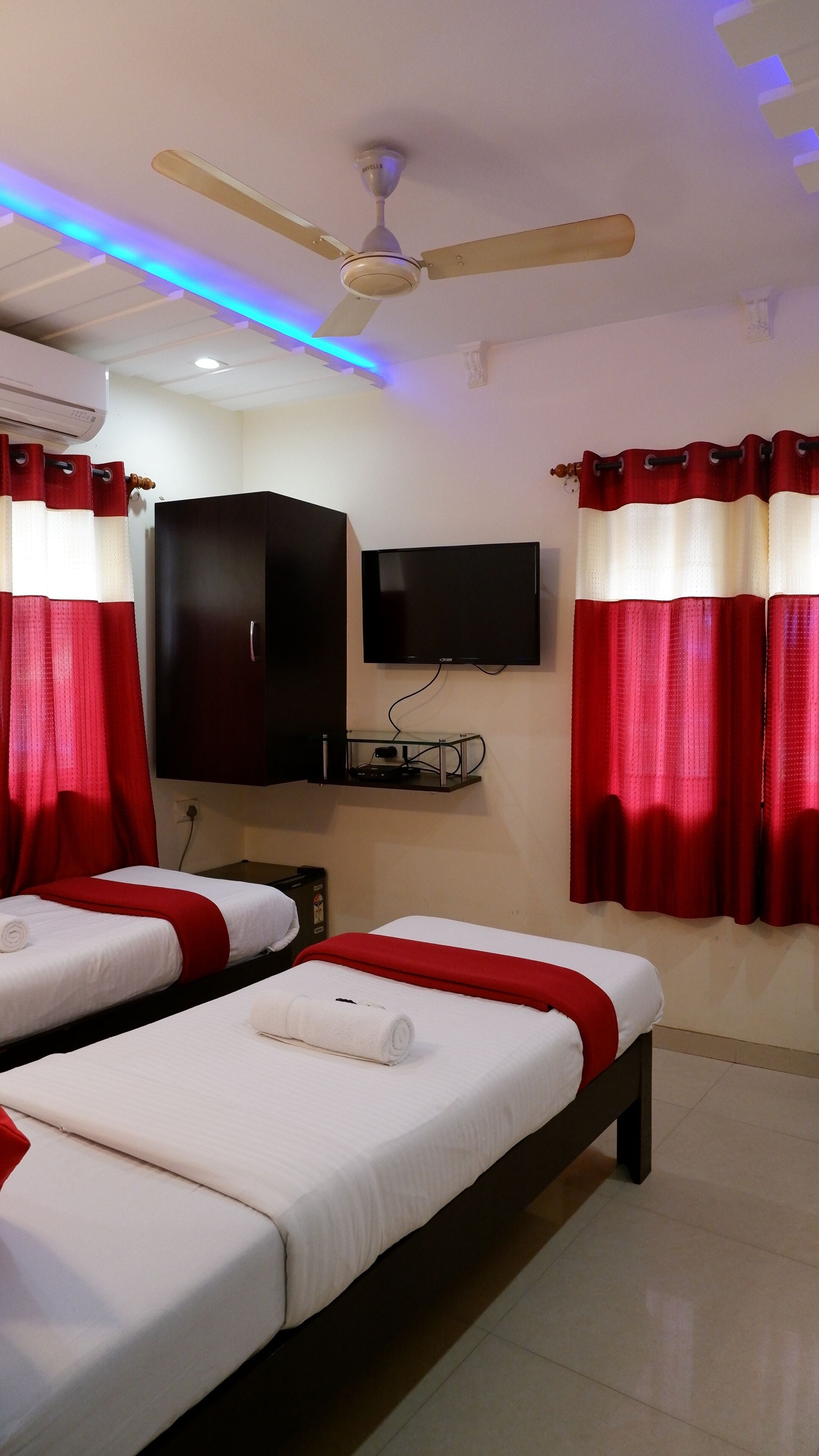 A hotel room with two beds, a ceiling fan, a mounted TV, and red curtains.