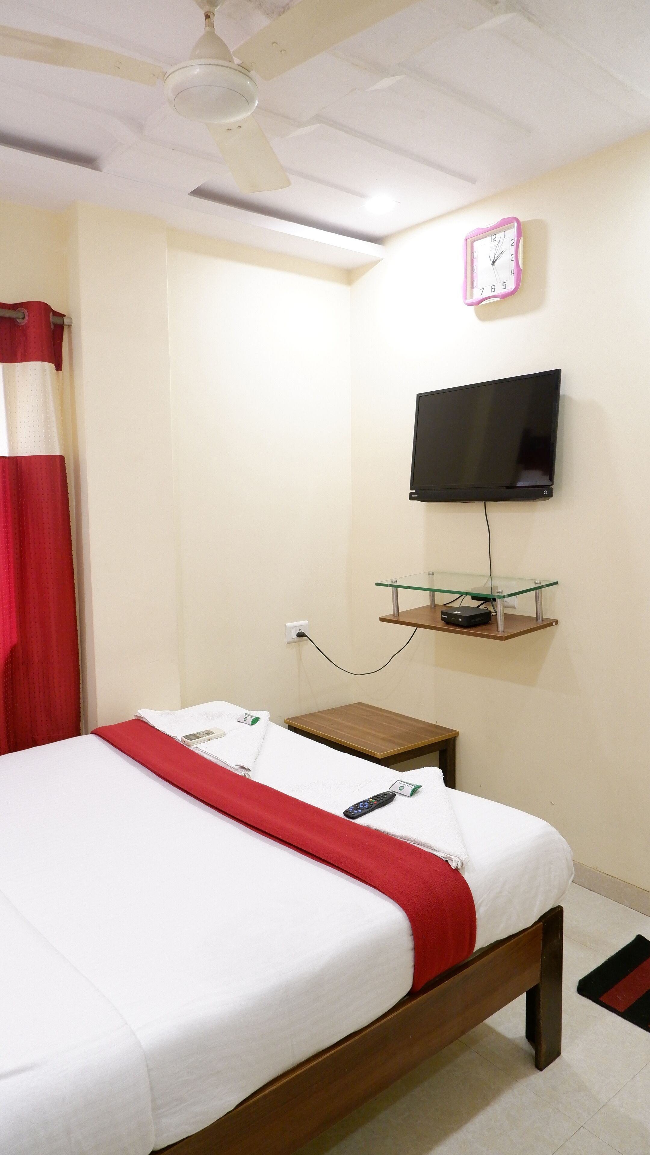 Una habitación de hotel con cama, televisor montado en la pared, mesita de noche y cortina roja.
