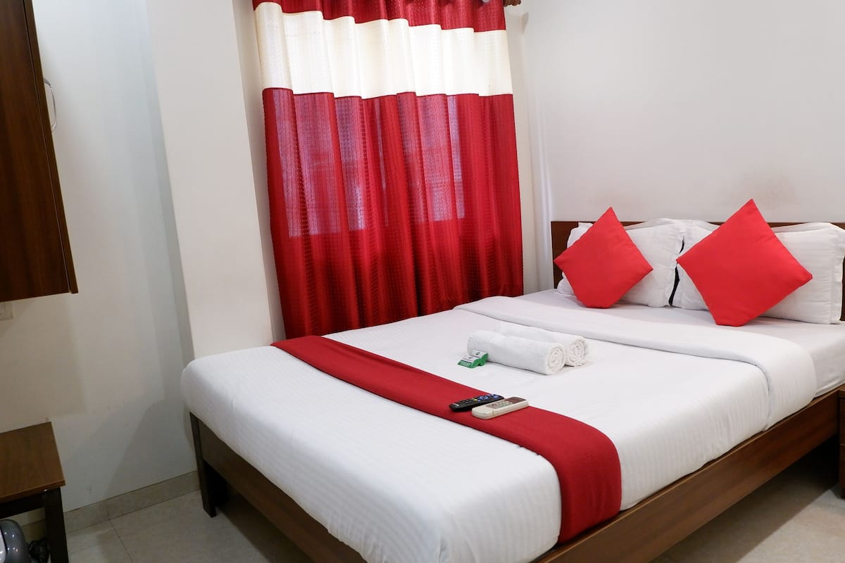 Una cama bien hecha con ropa de cama blanca y detalles rojos, una mesita de noche con un teléfono, y una cortina roja y blanca.