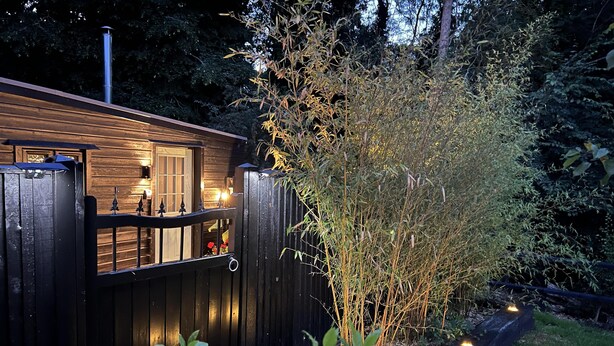 A wooden cabin with a balcony, surrounded by a garden with plants and a fence. - Charming Woodland Lodge 2 Person 1 Bedroom (Great Yarmouth)