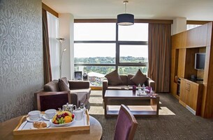 Habitación de hotel con dos sofás, una mesa de comedor con desayuno, televisor y un ventanal con vistas.