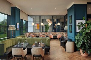 Un restaurante con zona de estar con asientos acolchados verdes, mesas de madera y lámparas colgantes.