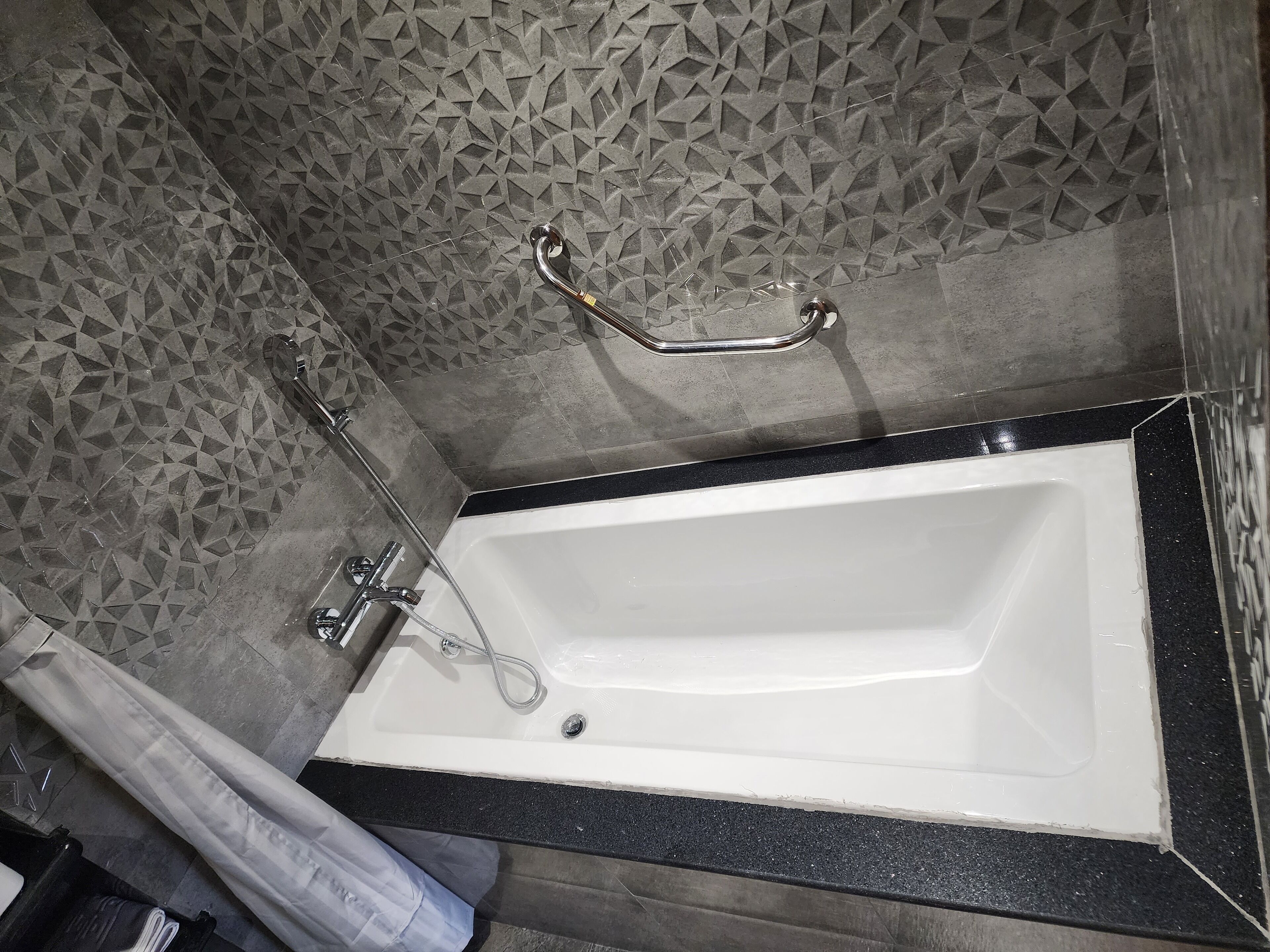 Una bañera con grifo de cromo, ducha y toallero, además de un revestimiento de pared texturizado.