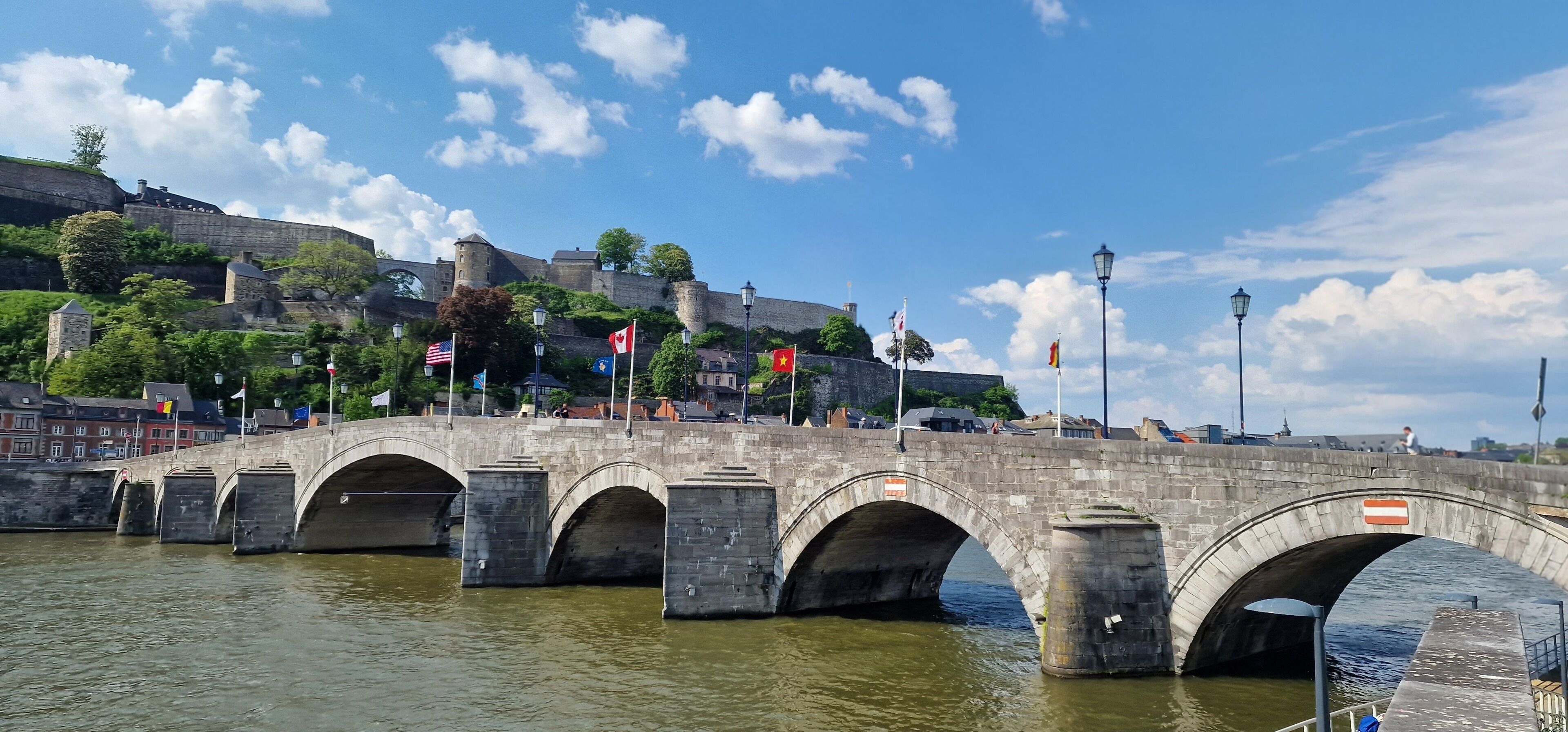 Un puente de piedra sobre un río con banderas, un castillo al fondo y un cielo despejado.
