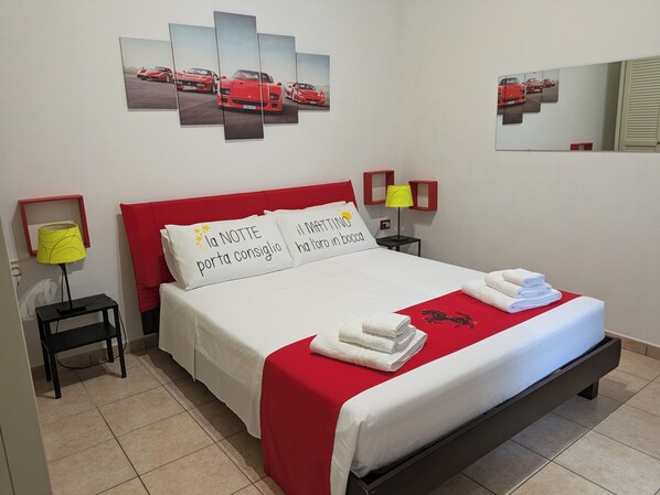 Ein ordentlich eingerichtetes Hotelzimmer mit einem Bett, Kissen, einem roten Kopfteil, einem Nachttisch mit Lampe und einem Wandbild mit Automotiv-Motiven.
