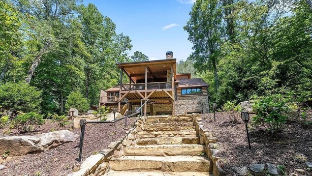 A log cabin with a stone staircase leading to a wooden deck, surrounded by dense forest. - On Toccoa River - Luxury Riverfront - Hot tub (Mineral Bluff)