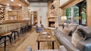 A rustic interior with a stone fireplace, leather sofa, wooden bar stools, and a cozy seating area. - On Toccoa River - Luxury Riverfront - Hot tub (Mineral Bluff)