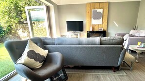 A modern living room with a grey sofa, a black armchair, a TV, a fireplace, and a view of greenery outside. - Twickenham Green (Twickenham)
