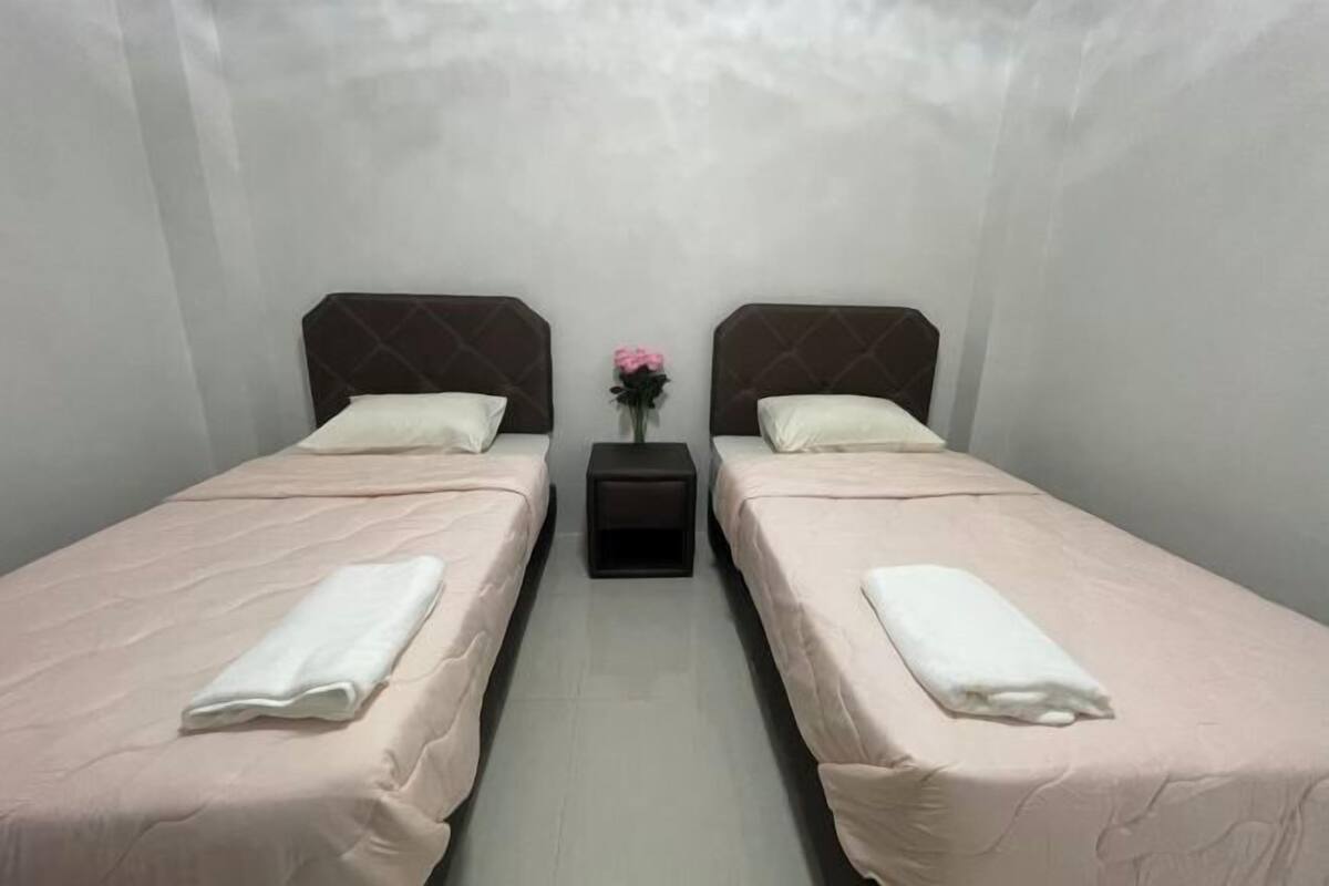A room with two beds, each with a pillow and a folded towel. There is a small table with a vase of flowers between the beds.