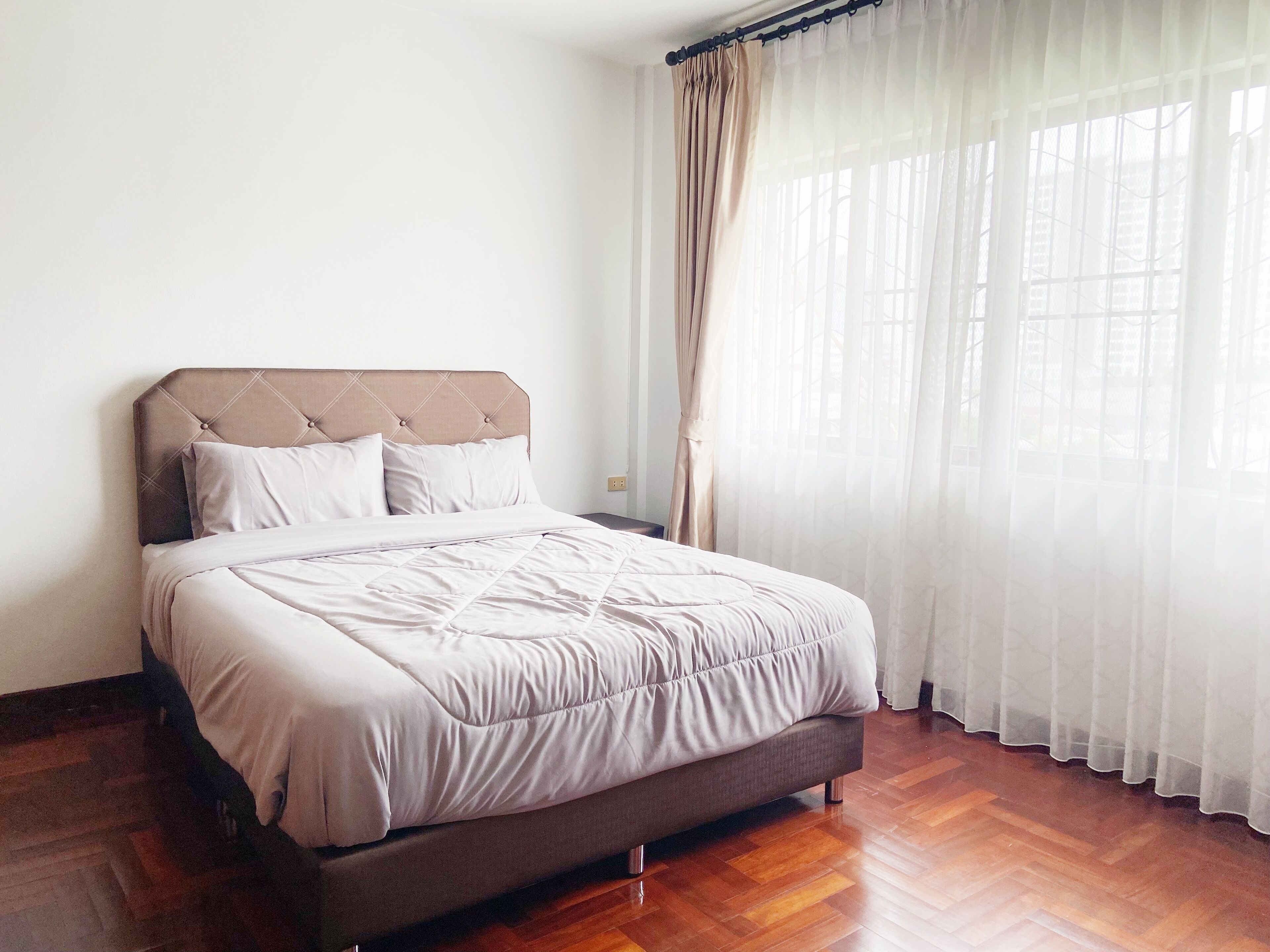 Una cama bien tendida con cabecera capitonada, piso de madera y cortinas traslúcidas en una ventana.