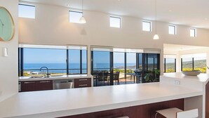 A modern kitchen with large windows overlooking the sea, a white countertop, and bar stools.
