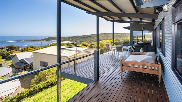 A balcony with a bed, a table, and a chair, overlooking a coastal landscape.